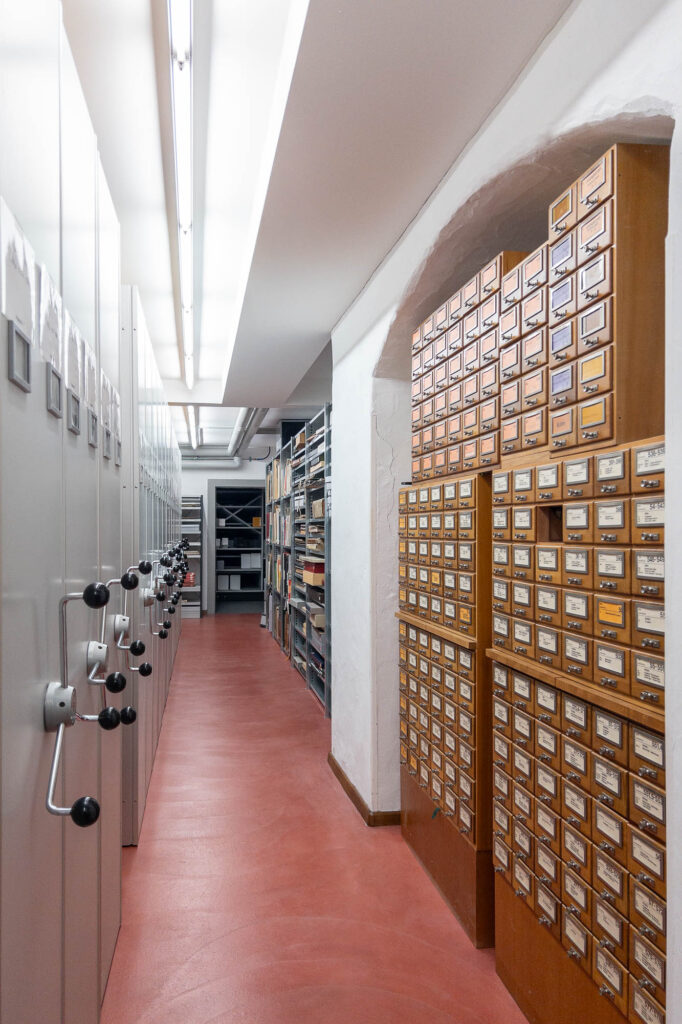 A narrow corridor with red flooring lined with wood card catalogue cabinets on one side and mobile shelving units with hand cranks on the other.