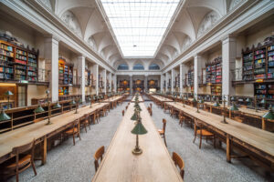 A large, bright library reading room with rows of wooden tables and chairs, green desk lamps, tall bookshelves filled with books, and a high arched ceiling with skylights.
