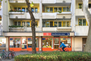 A cyclist rides past the entrance of a library marked with a sign reading “buechereien wien", located on the ground floor of a residential building with balconies lined with plants and yellow railings.