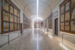 A long, arched hallway with polished stone floors and framed plaques on both walls, separated by thin railings; light enters from a doorway at the far end of the corridor.