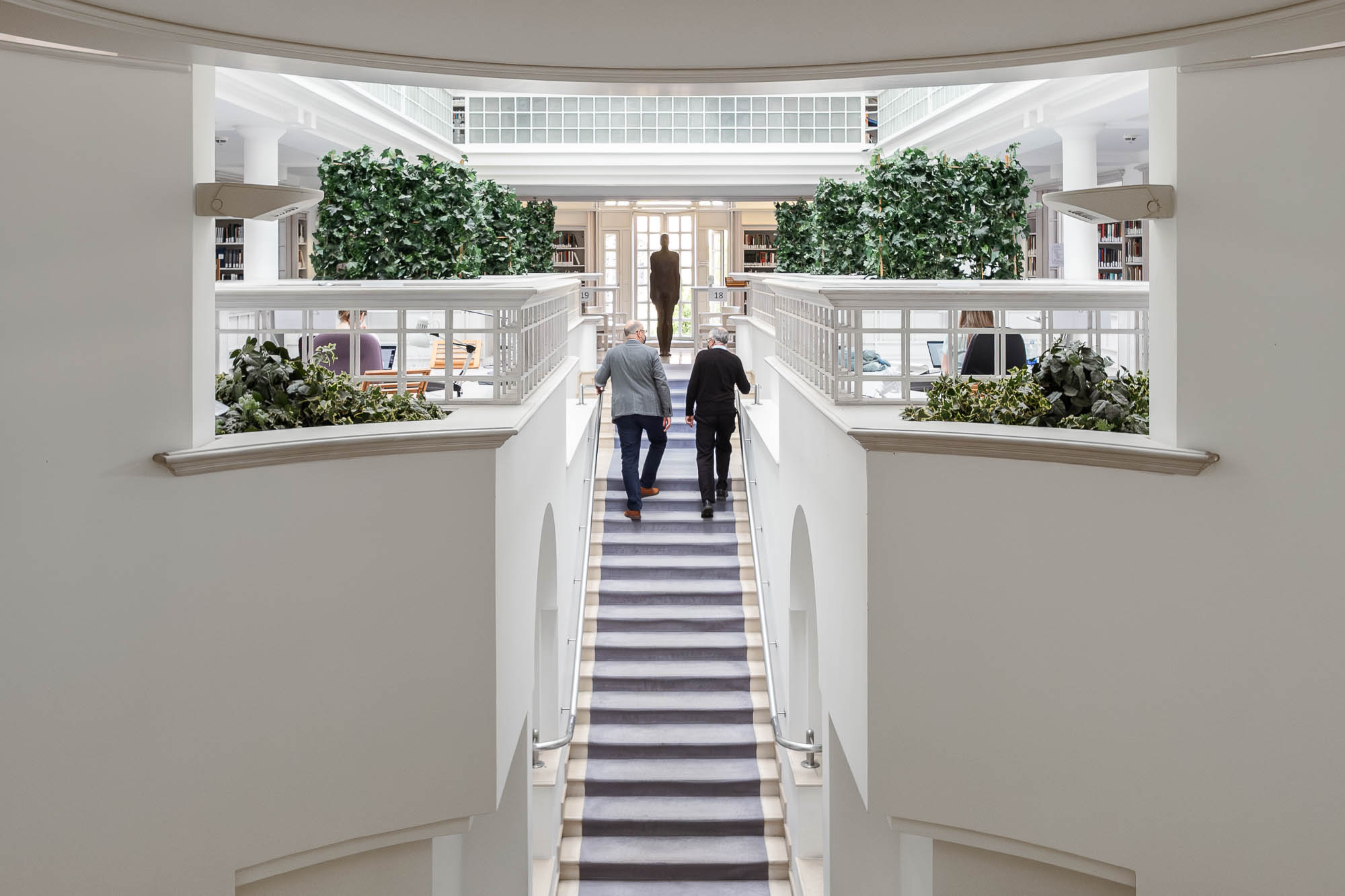Two people walk up a wide staircase in a bright, modern building with white walls, indoor plants, and large windows. A statue stands at the top of the stairs, and bookshelves are visible in the background.