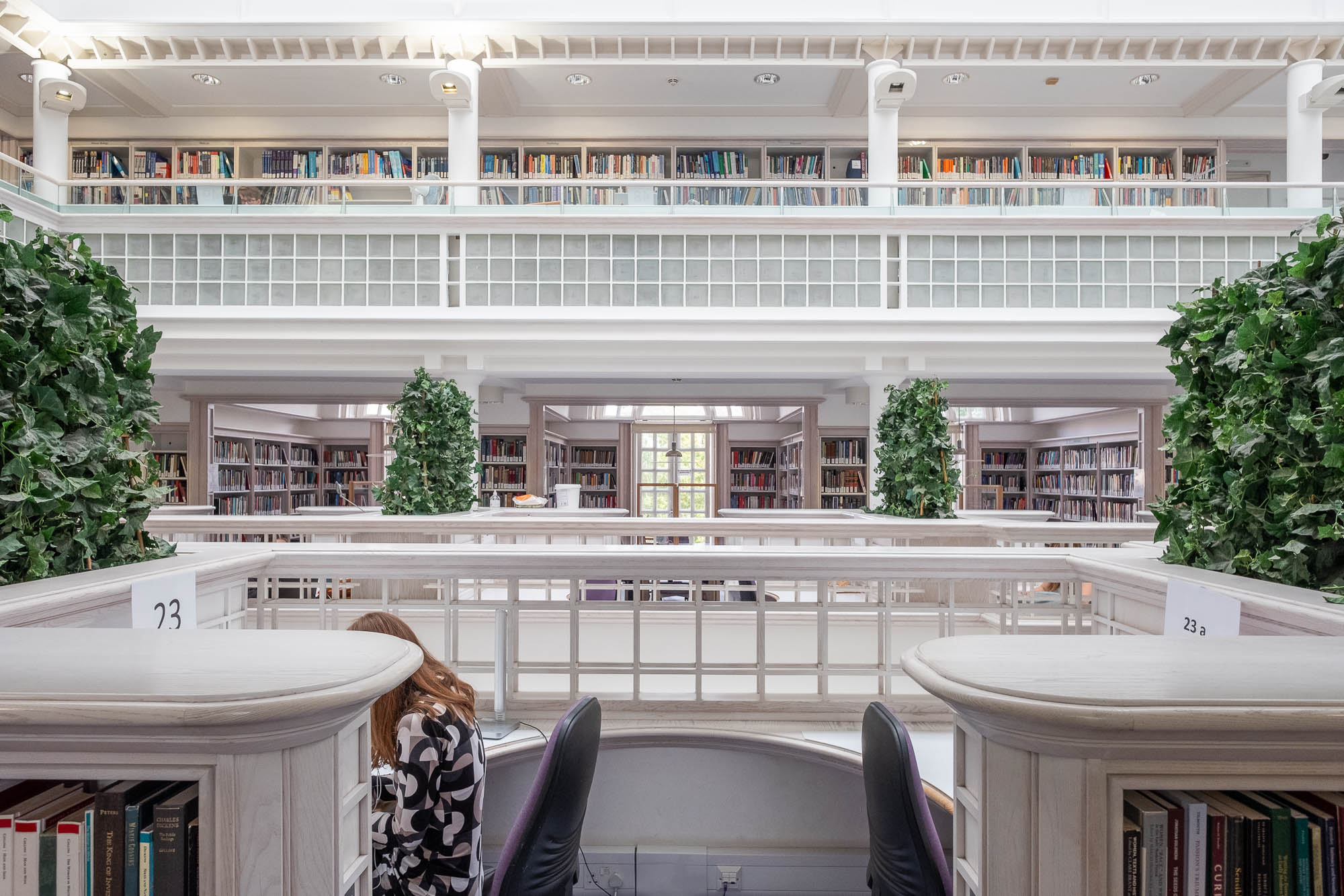 A bright library interior with bookshelves on two floors, white railings, green potted plants, and a person seated at a study desk, partially visible in the lower left corner.