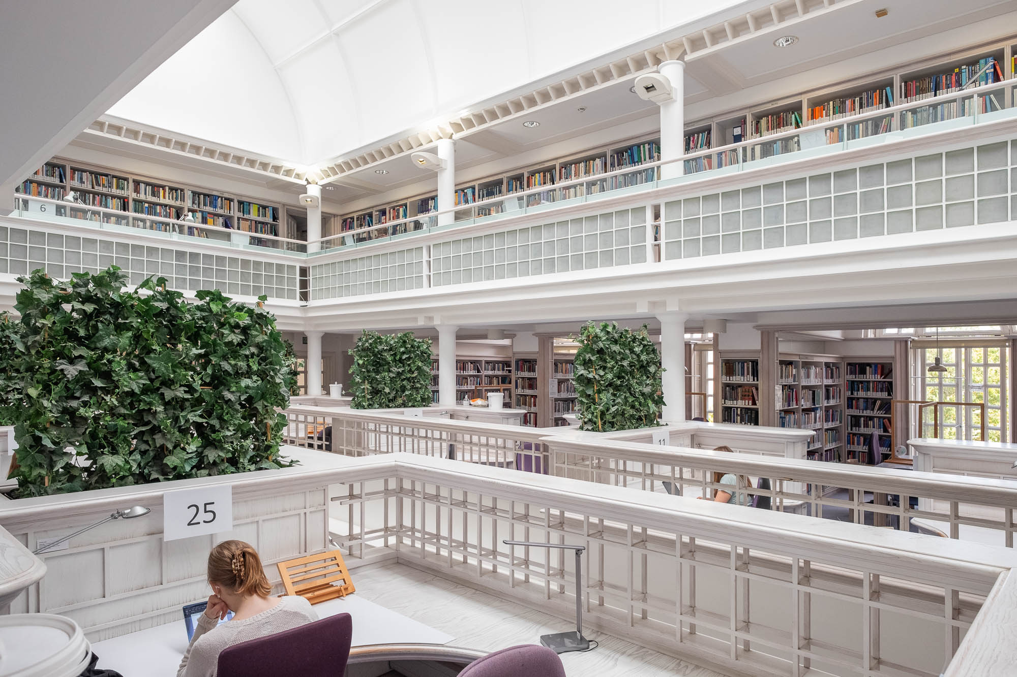 A spacious, modern library interior features tall bookshelves on two levels, large green plants, white railings, and several people studying at desks. Natural light enters through a high, arched ceiling.