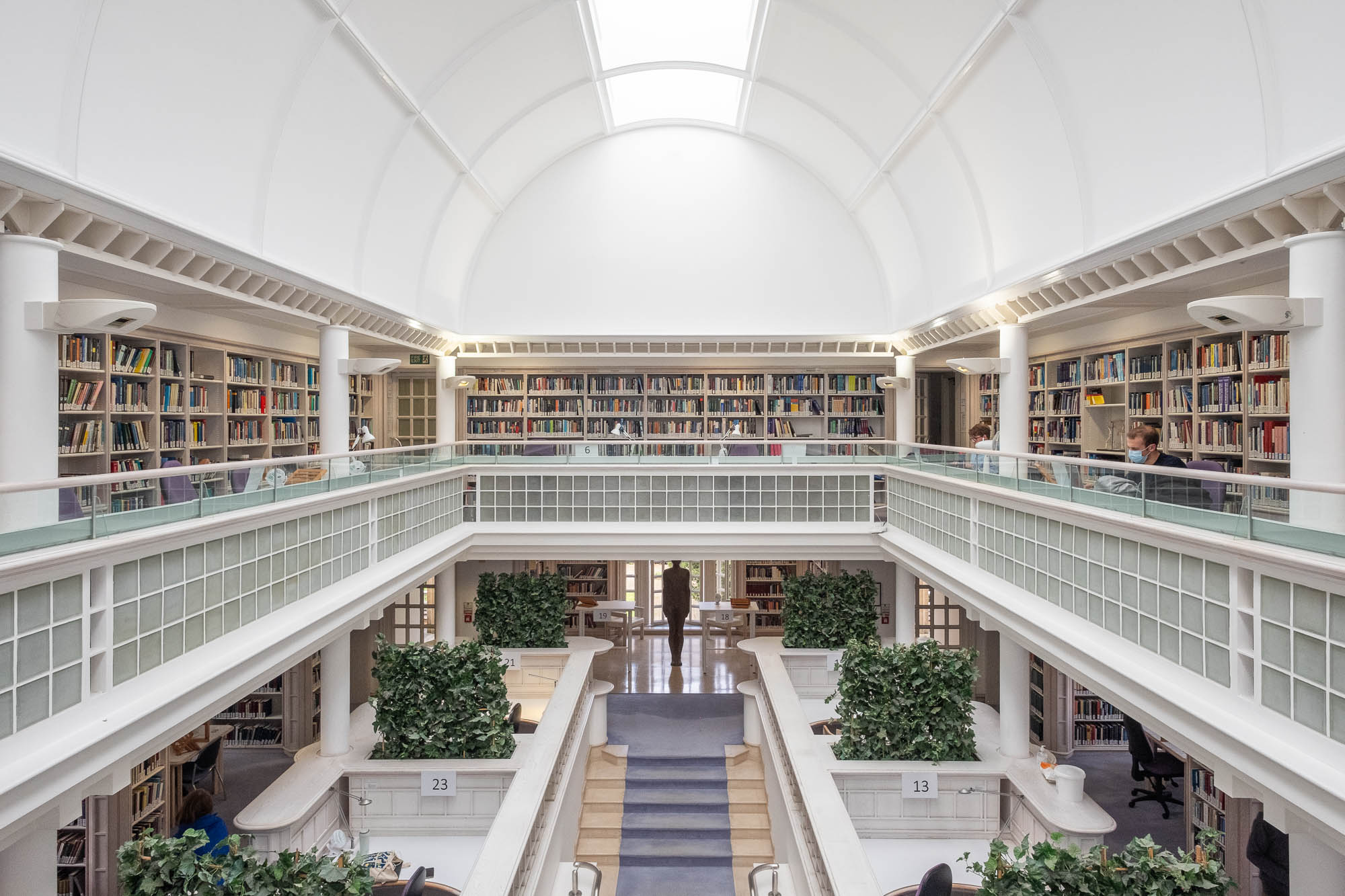 A spacious, bright library with two levels of bookshelves, white walls, a high arched ceiling, and glass barriers. People are reading and working at desks on a mezzanine. Green plants decorate study areas on the lower floor.
