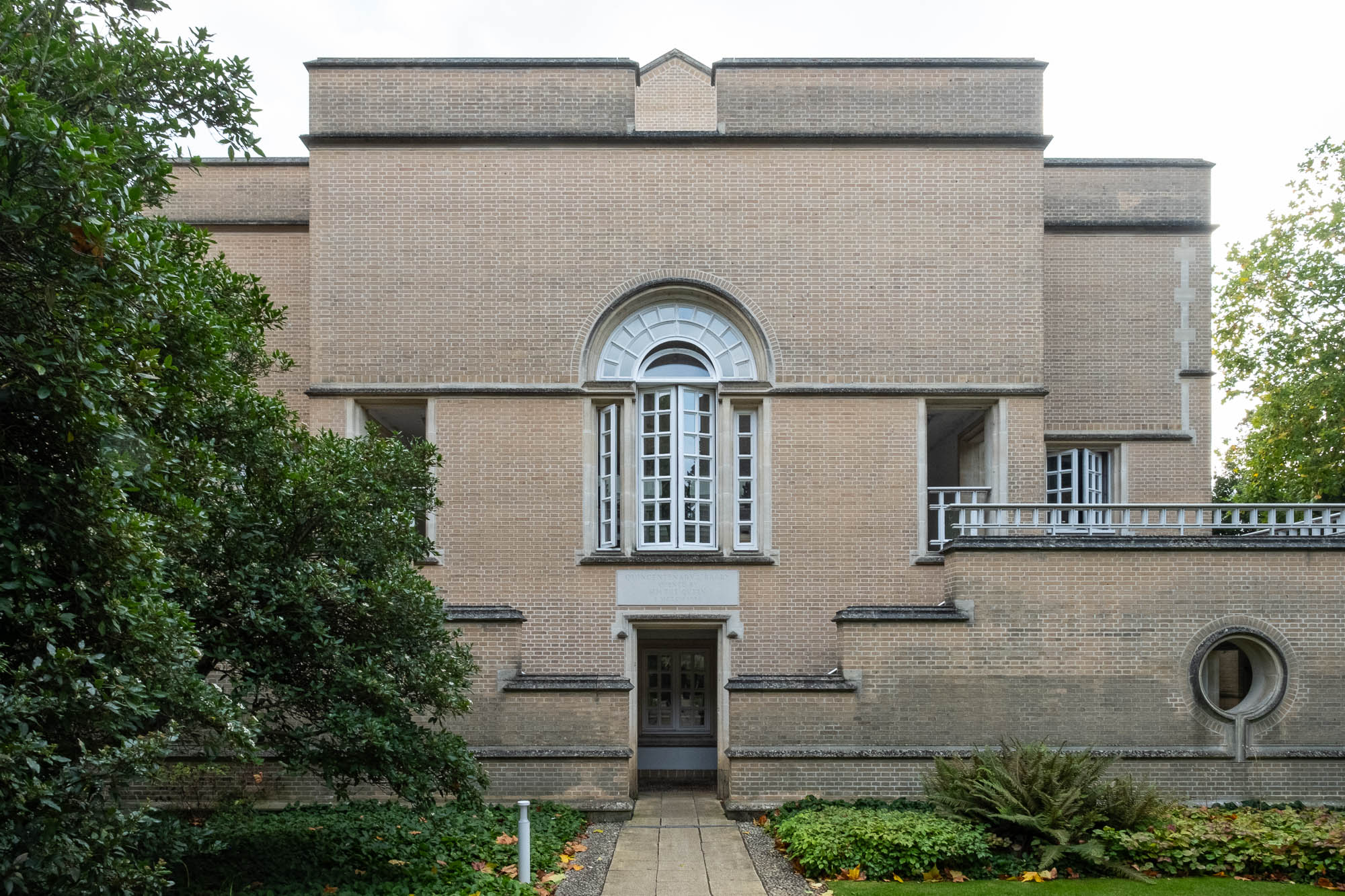 A symmetrical brick building with geometric shapes features a large arched window above a smaller entrance door, rectangular and circular window details, and landscaped greenery on either side of the stone path.