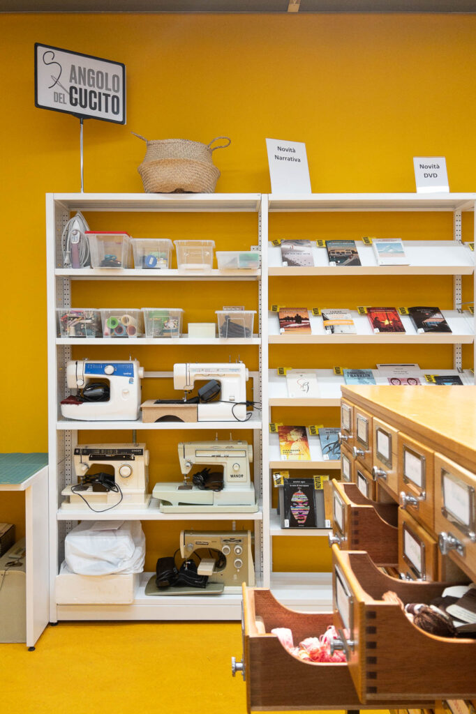 A white shelving unit against a yellow wall displays sewing machines, baskets, and books. Card catalog drawers are open in the foreground. Signs on the shelves read "Angolo del cucito", "Novità Narrativa" and "Novità DVD".