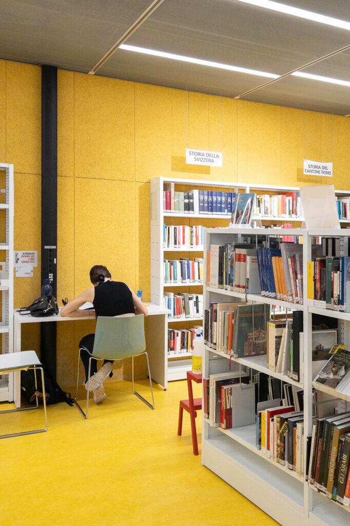 A person sits alone at a desk with books and papers in a library, surrounded by bookshelves labeled for different sections against yellow walls and flooring. Overhead lights illuminate the space.