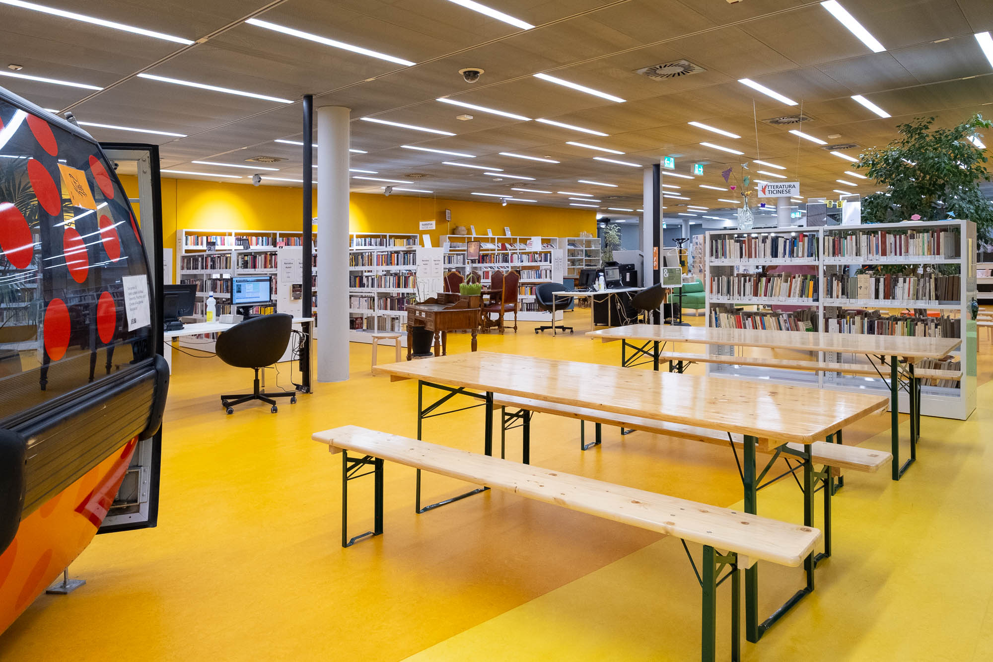 A modern library interior with wooden tables and benches in the foreground, bookshelves filled with books, computers, and chairs in the background, and yellow flooring and walls. The space is well-lit with ceiling lights.
