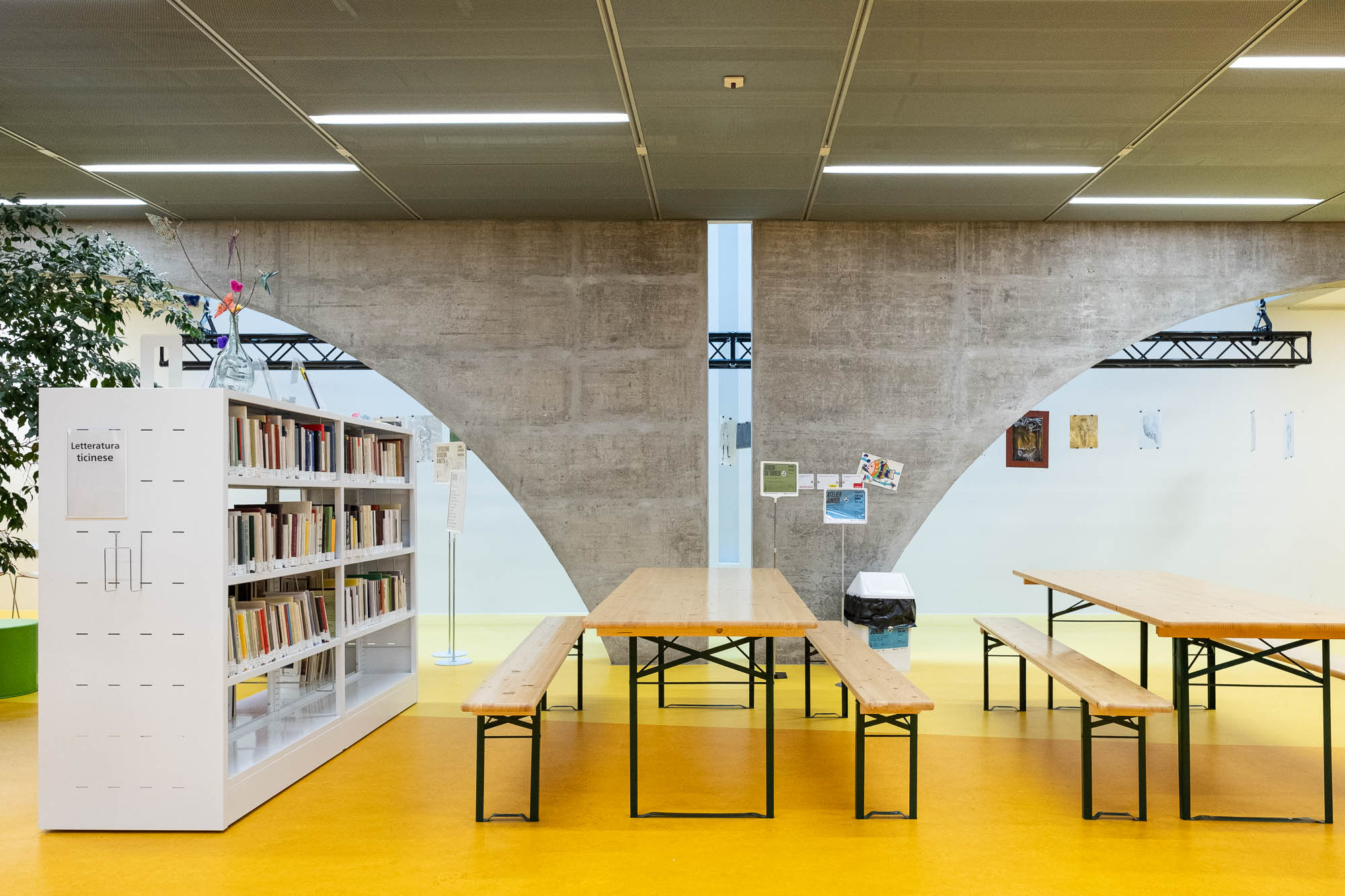 A library space with a white bookshelf filled with books on the left, wooden tables and benches in the center, a yellow floor, and a concrete wall with curved arches in the background. Artworks are displayed on the right wall.