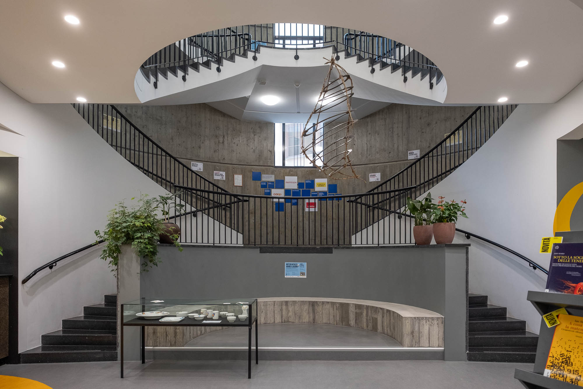 A modern interior with a curved staircase, circular opening above, potted plants on both sides, a glass display table, and a bench in the center. Blue and yellow papers are displayed on the back wall.