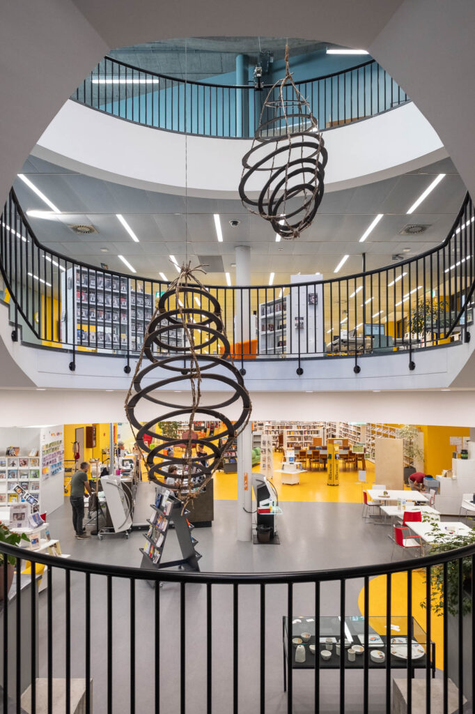 A modern library interior with two floors, circular railings, spiral wooden light fixtures, bookshelves, tables, and people using the space. The ground floor has displays and scattered books.