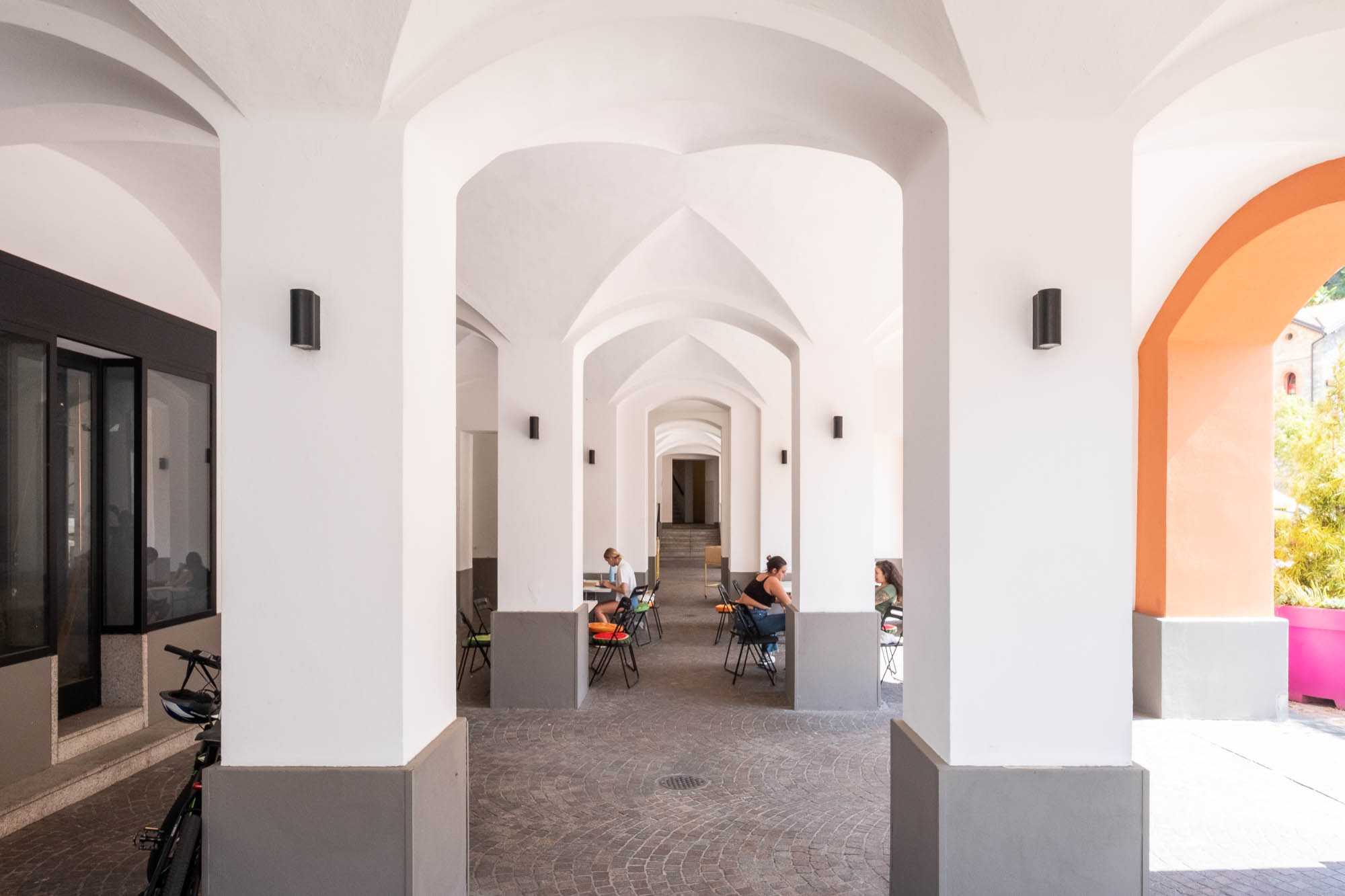 A covered outdoor walkway with white arched columns, stone flooring, and people sitting at tables on either side. There are modern wall lights and a bicycle parked on the left. Stairs are visible at the far end.