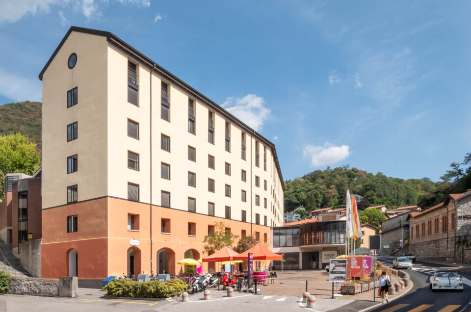 A large, light yellow and orange building with multiple floors stands at the center, featuring a courtyard with outdoor seating, umbrellas, and parked motorcycles; hills and clear sky are visible in the background.