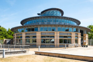 A round, modern building with large windows and brick accents, set on a paved area with a sign and some greenery in the background under a clear blue sky.