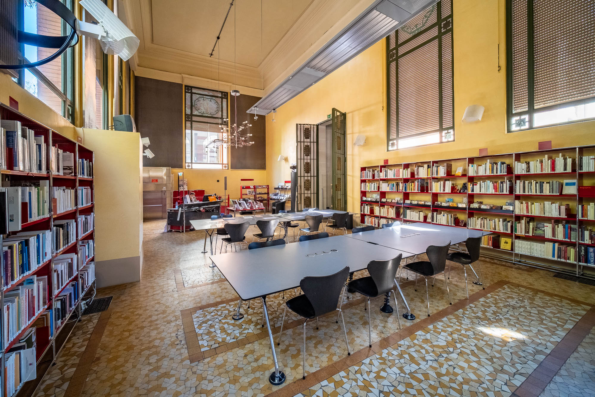 A spacious library room with tall ceilings, yellow walls, shelves filled with books, large windows with blinds, and two rectangular tables surrounded by black chairs on a patterned tile floor.