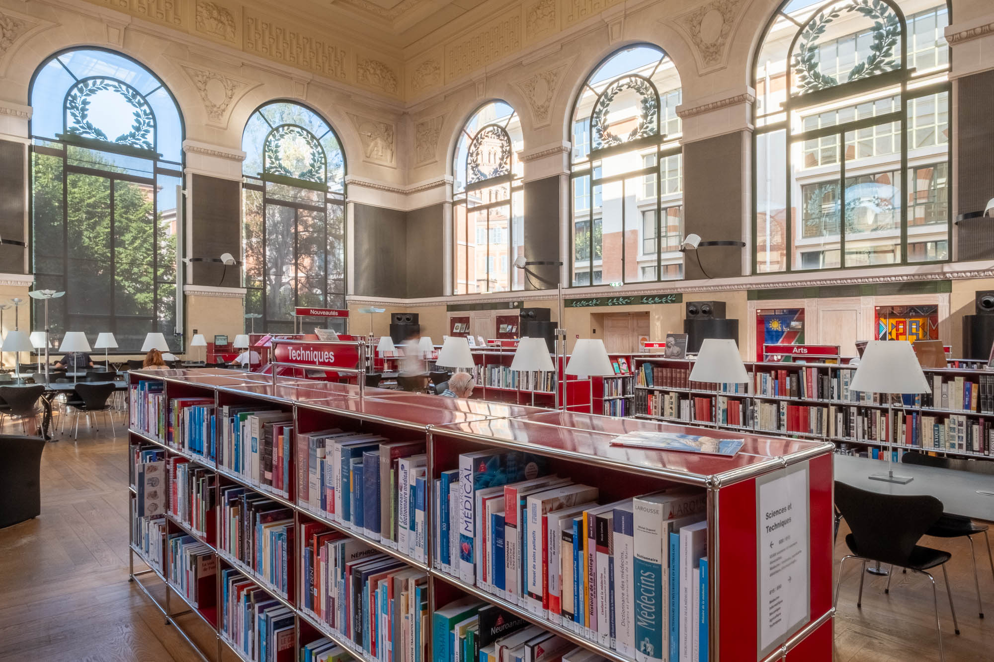 Spacious library interior with tall arched windows, wooden floors, red bookshelves filled with books, and white reading lamps on tables; sunlight streams through the windows.
