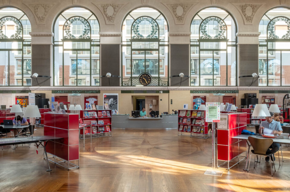 A spacious library interior with large arched windows, wooden floors, red bookshelves, a central information desk, a clock on the back wall, and several people reading or working at tables.