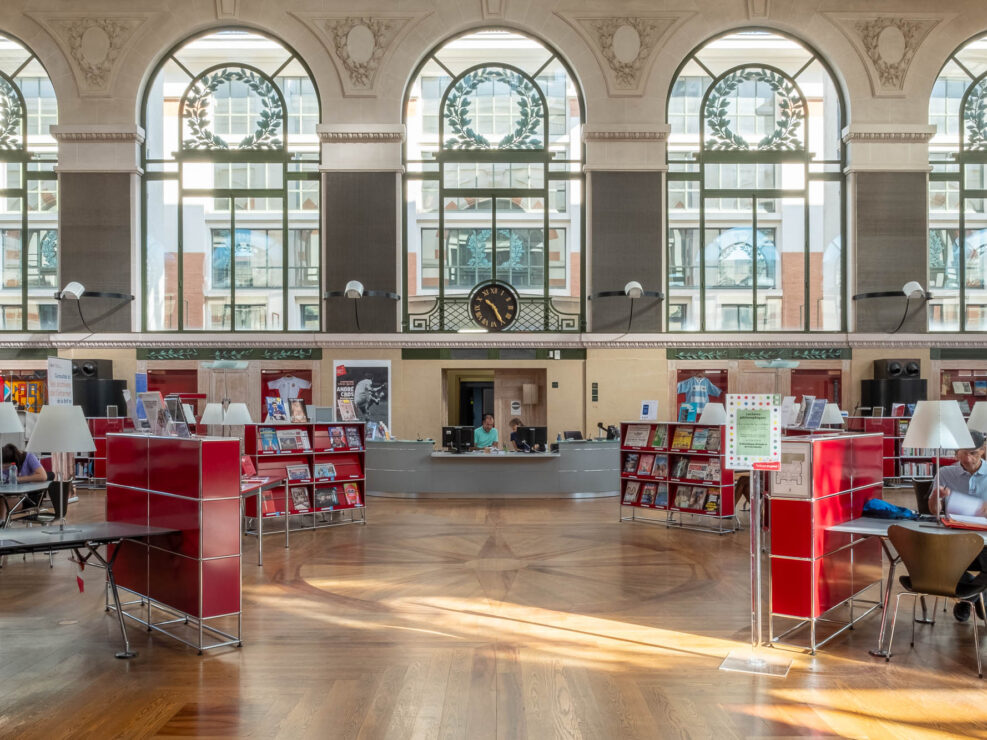 A spacious library interior with large arched windows, wooden floors, red bookshelves, a central information desk, a clock on the back wall, and several people reading or working at tables.