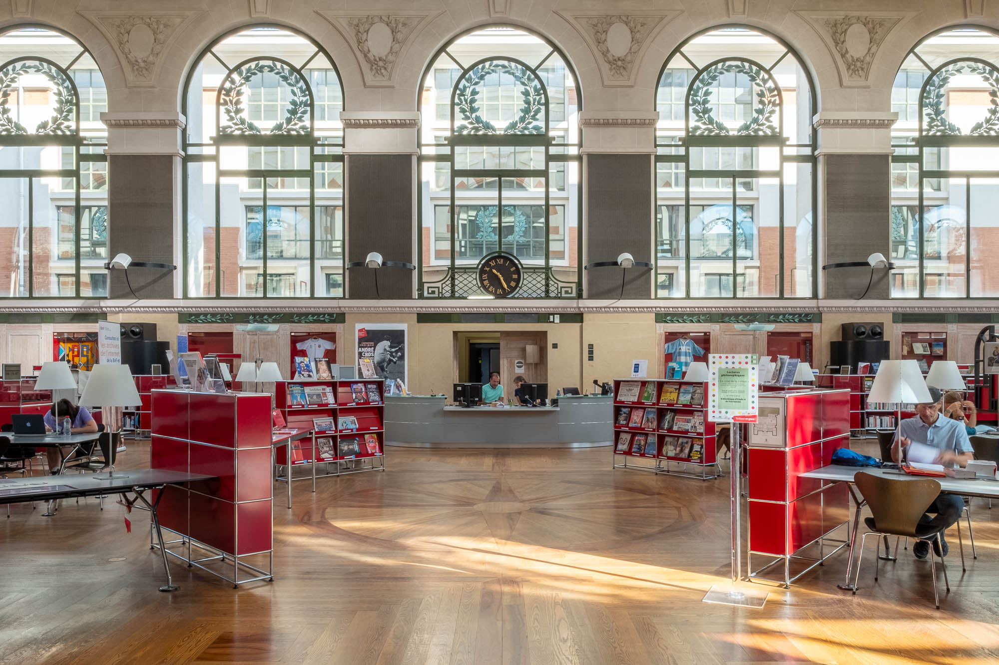 A spacious library interior with large arched windows, wooden floors, red bookshelves, a central information desk, a clock on the back wall, and several people reading or working at tables.