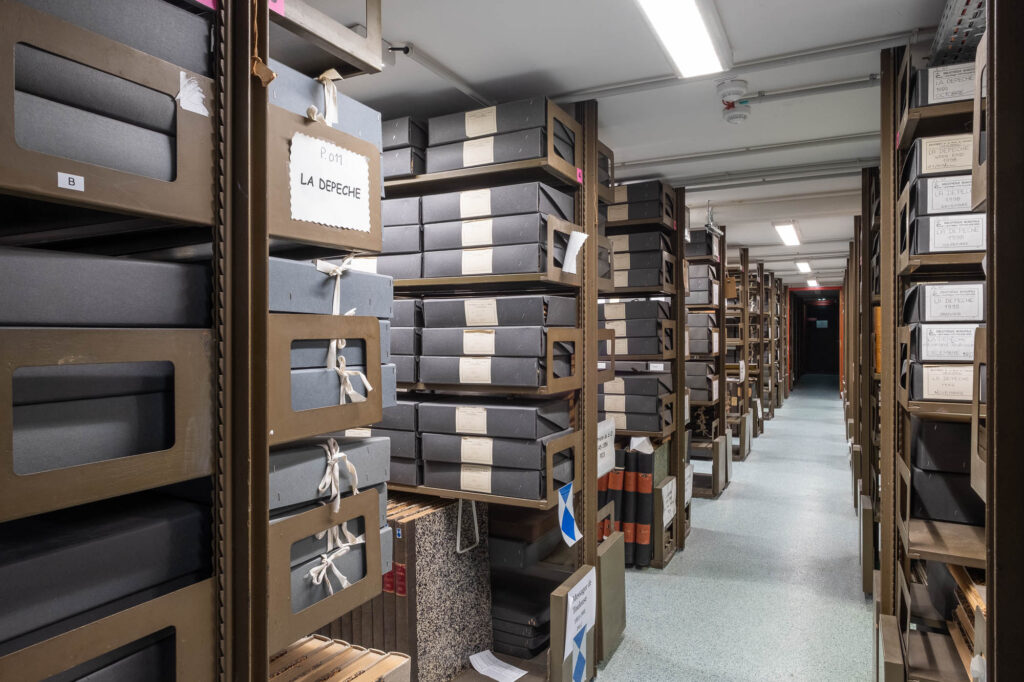 Rows of shelves filled with labeled archival boxes and folders in a well-lit storage room, with signs and tags attached to some boxes, and a long hallway extending into the distance.