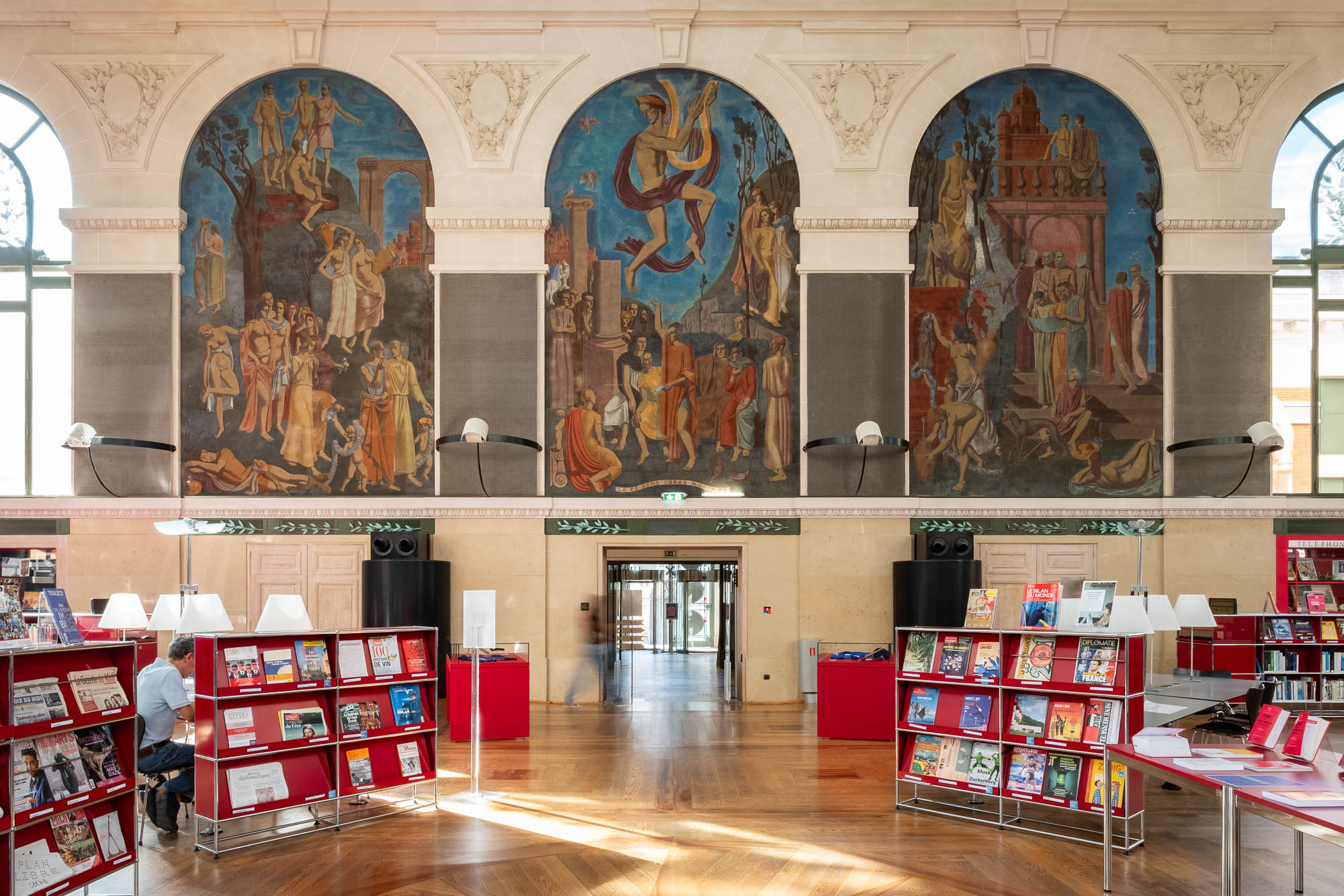 A spacious library interior with wooden floors, red bookshelves displaying books and magazines, tall arched windows, and large wall murals depicting classical scenes above the shelves.