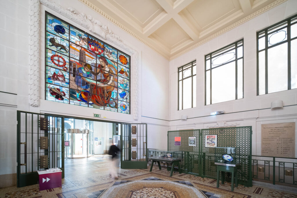 A spacious, bright interior features large windows, a decorative mosaic floor, green metal gates, and a colorful stained glass window depicting workers. A person walks near museum displays and informational panels.