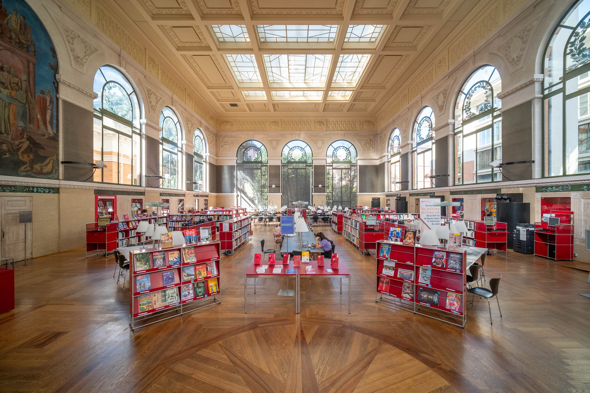Large, bright library with high ceilings, tall arched windows, red book displays, and wooden floors. Books and magazines are organized on shelves and tables; a few people are seen reading or browsing.