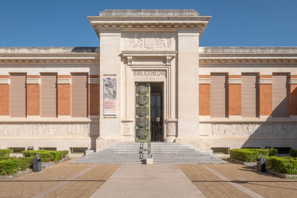 A symmetrical facade of a neoclassical building labeled BIBLIOTHEQUE above the entrance, with red brick details, sculptures, and a staircase leading to glass doors, surrounded by trimmed bushes and a paved walkway.