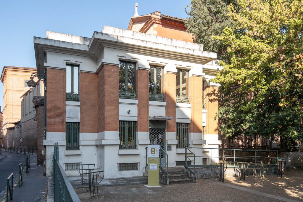 A two-story building with red brick and white walls, tall vertical windows with bars, and a sign near the entrance. There is a tree to the right and a narrow street to the left.