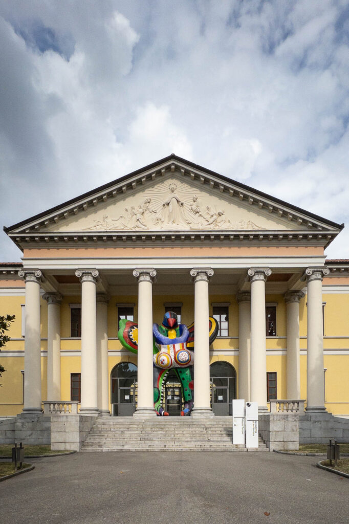 A neoclassical building with yellow walls and tall white columns features a large, colorful abstract sculpture displayed at the entrance. The sky above is partly cloudy.