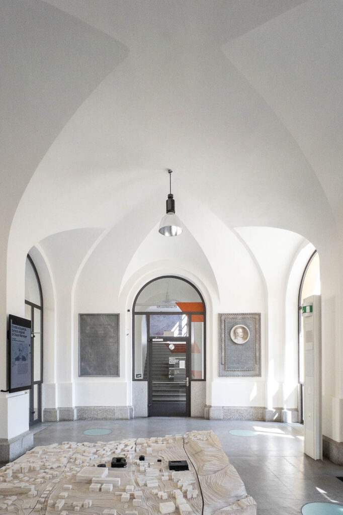 A bright, vaulted hallway with arched doorways and a central hanging light fixture. A large architectural model is displayed on a table in the foreground. A door and wall plaques are visible in the background.