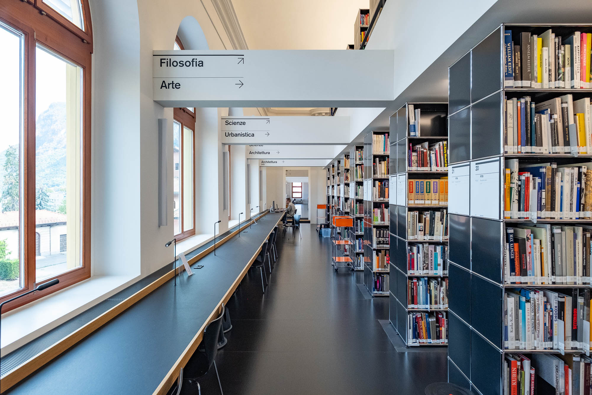 A modern library interior with long study tables, black chairs, tall bookshelves filled with books, large arched windows, and overhead directional signs in Italian indicating sections like "Filosofia", "Arte", and "Scienze".