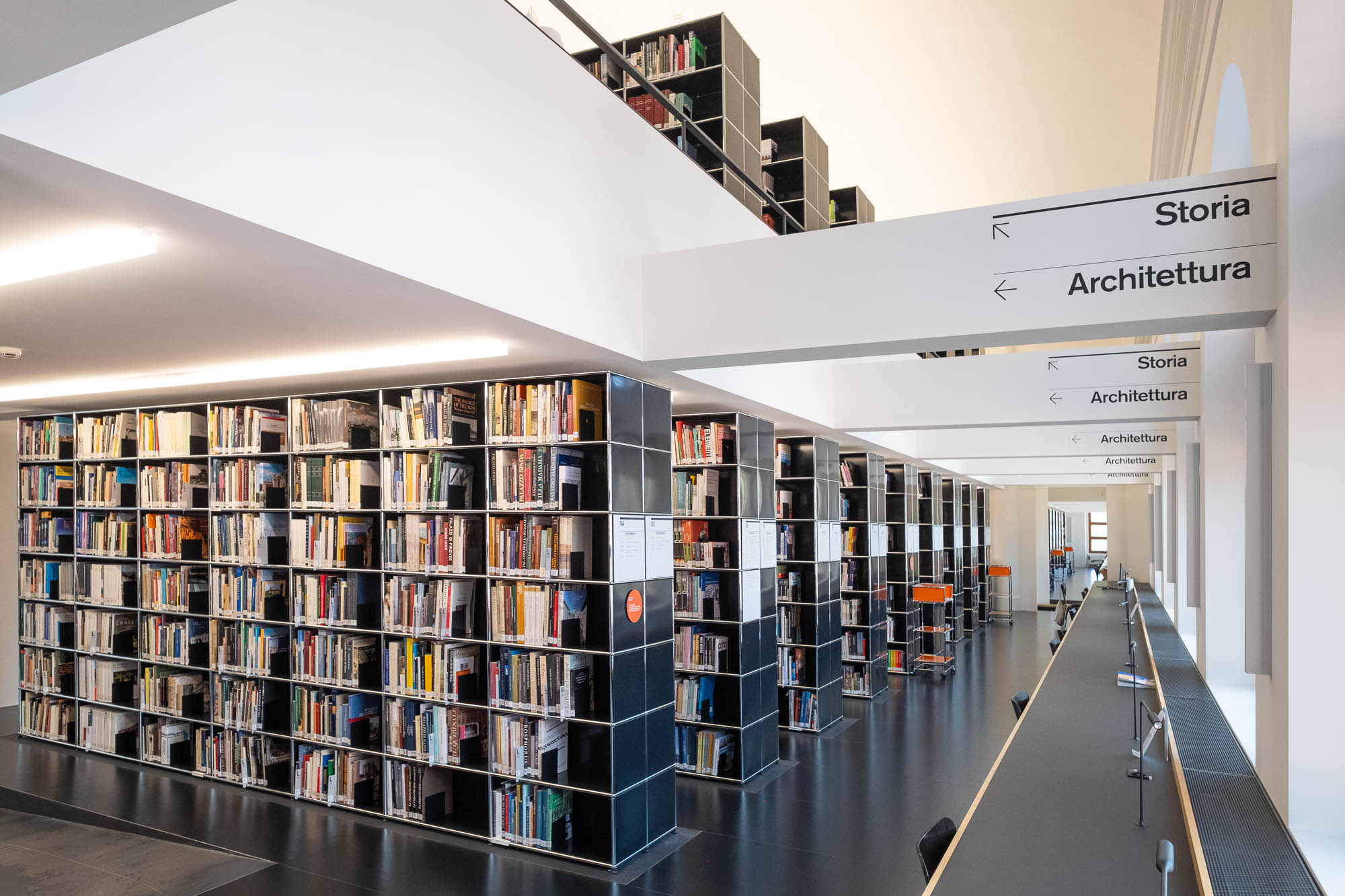 A modern library interior with tall bookshelves filled with books, sign decals reading "Storia" and "Architettura", and long study tables with chairs along large windows.