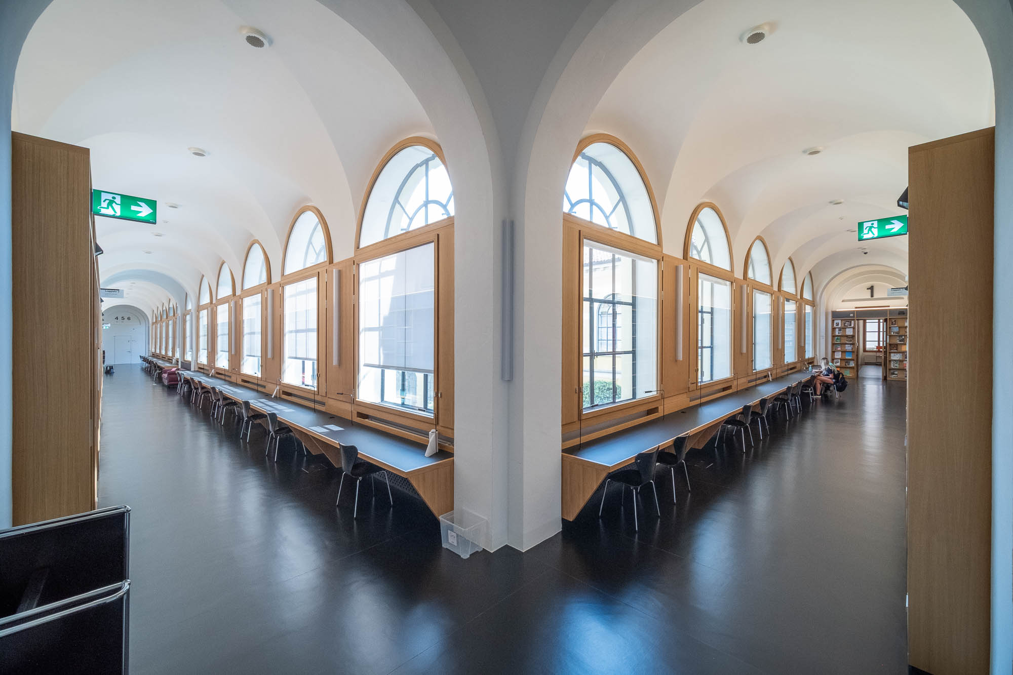 A spacious, well-lit library study area with long rows of desks and chairs beneath large arched windows. The floor is dark and shiny, and there are exit signs on the ceiling. Shelves of books are visible at the far end.