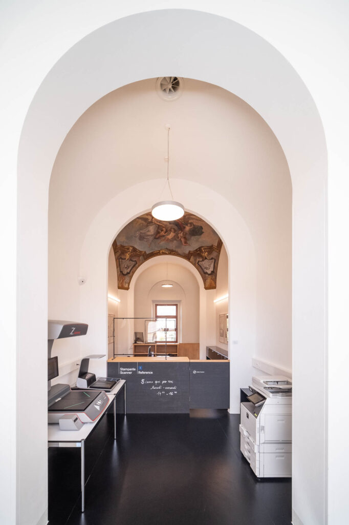 A library copy area with scanning and printing equipment, white walls, a black floor, and an arched ceiling featuring a classical mural. A circular light hangs from the ceiling. Natural light comes through a window at the back.