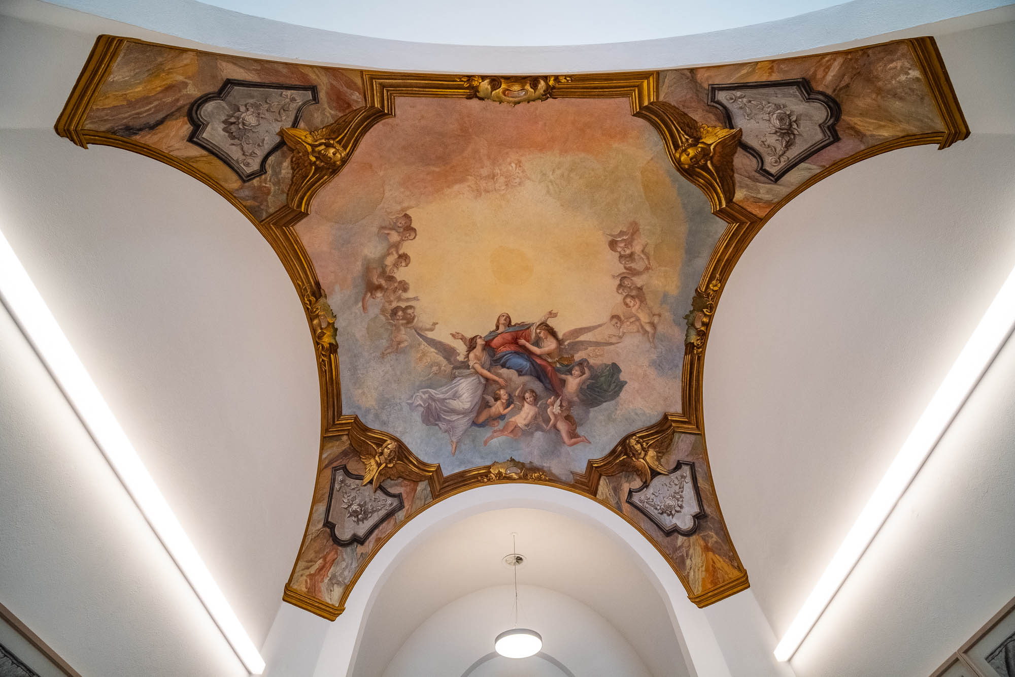 A ceiling fresco featuring several figures in flowing robes, surrounded by clouds and framed by ornate golden architectural details, with two fluorescent lights on either side of the hall below.