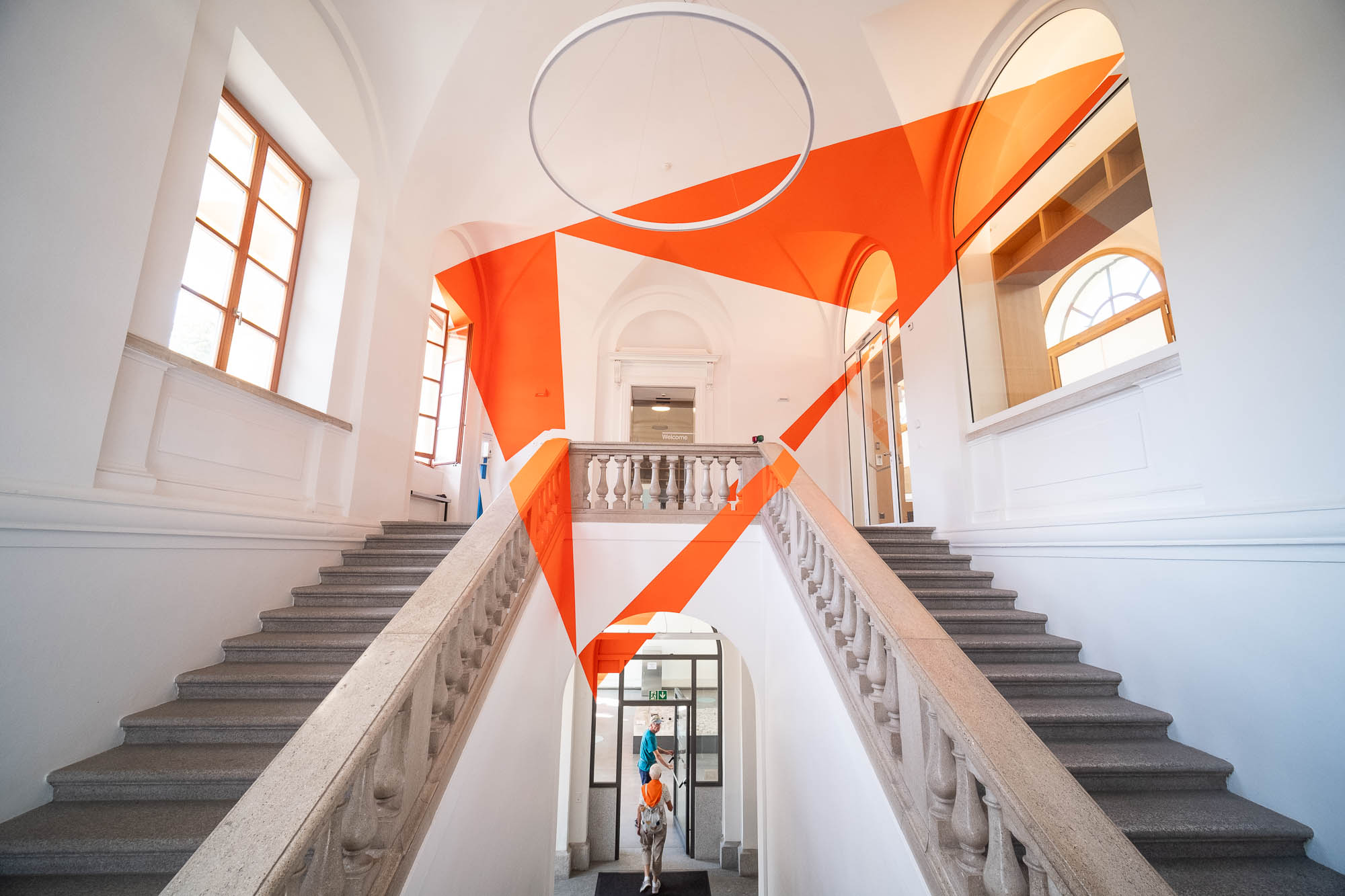 Two people are exiting through a door at the bottom of a split staircase in a bright, white interior with tall arched windows. An orange geometric art installation is painted on the ceiling above the stairs.