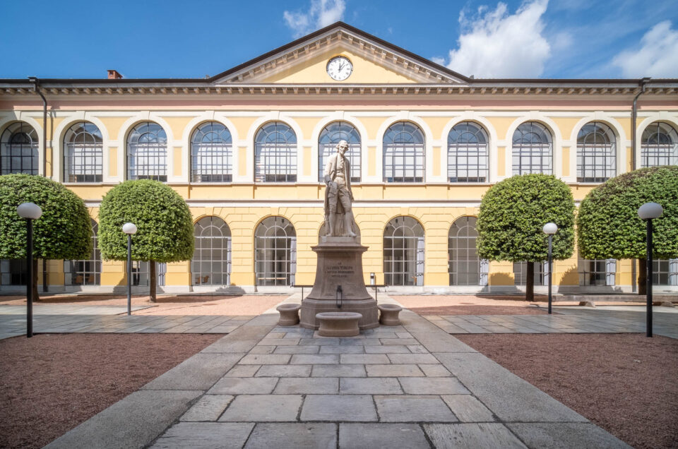 A symmetrical courtyard with a central statue and fountain, surrounded by neatly trimmed trees, leads to a yellow two-story building with arched windows, a clock, and a gabled roof under a partly cloudy sky.