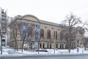 A large historic building with arched windows and columns stands on a snowy street. Leafless trees line the sidewalk, and blue banners hang from the facade. Snow covers the ground and parts of the building.