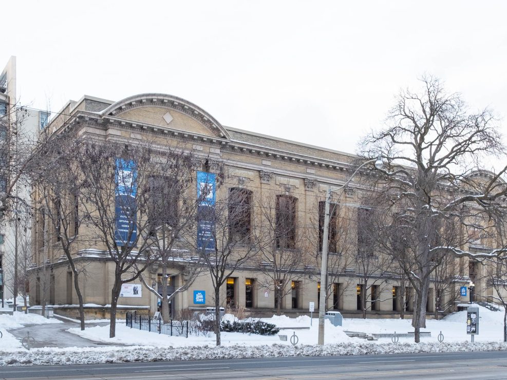 A large historic building with arched windows and columns stands on a snowy street. Leafless trees line the sidewalk, and blue banners hang from the facade. Snow covers the ground and parts of the building.