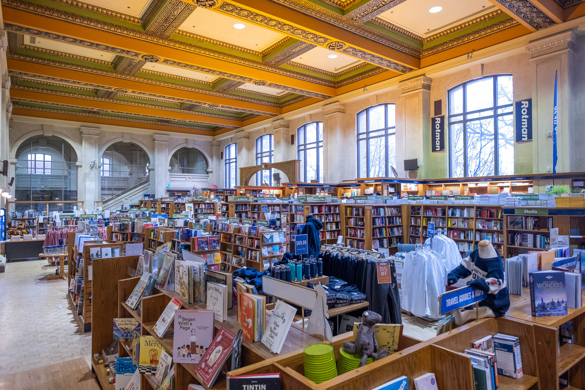 A large, brightly lit bookstore with tall arched windows, colorful decorative ceiling, and rows of bookshelves and display tables filled with books, clothing, and gifts. Signs categorize sections, and natural light fills the space.