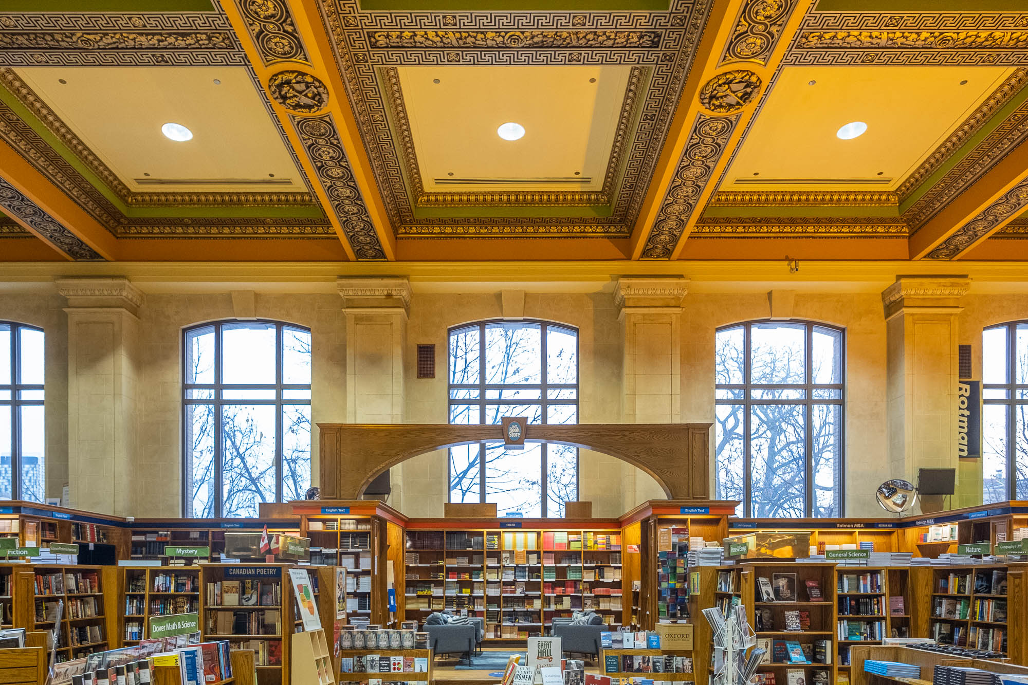 A spacious library interior with tall windows, ornate coffered ceiling, and bookshelves filled with books. Two desks are positioned near the center, and natural light enters through the large windows.