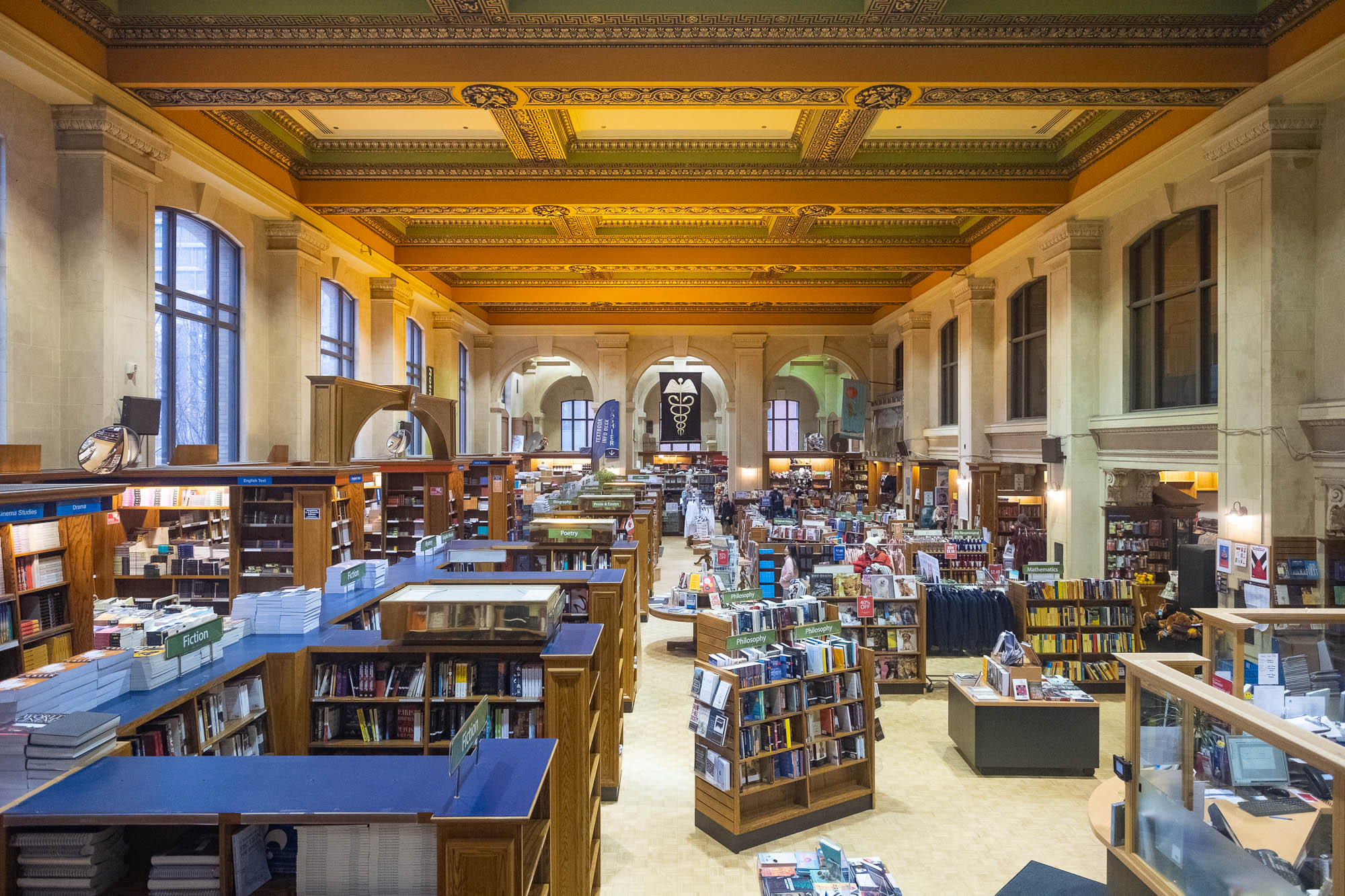 A spacious library interior with tall bookshelves filled with books, tables, and display stands under a decorative, ornate ceiling with green and gold designs. Large windows allow natural light into the room.