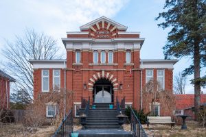 A two-story red brick building with white trim and arched entrance, marked Carnegie A.D. 1909 above the door. Steps lead to the entrance, flanked by leafless trees. The sky is partly cloudy.