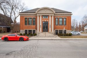 A red sports car is parked in front of a symmetrical brick building with columns and arched architectural details on a cloudy day. The street and surrounding area are quiet.