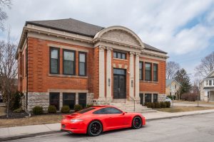 A red sports car is parked in front of a symmetrical brick building with columns and arched architectural details on a cloudy day. The street and surrounding area are quiet.