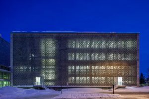 A modern building with a patterned metal facade is lit from within at night. Snow covers the ground, and two lit entrances are visible on either side of the structure.