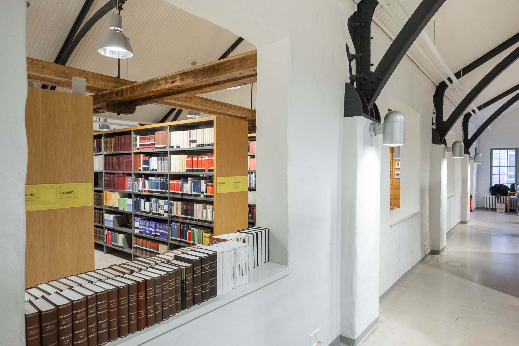 A modern library interior with bookshelves filled with books, a row of books on a ledge, white walls, exposed beams, and industrial-style lighting fixtures. A window on the right lets in natural light.