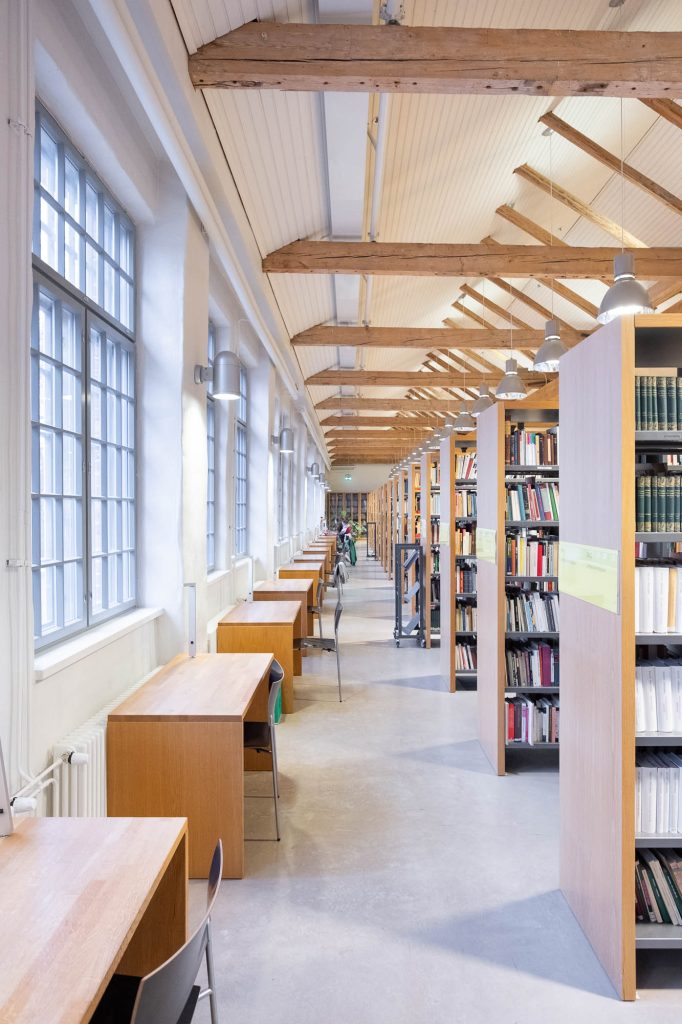 A bright library interior with tall bookshelves on the right and rows of wooden study desks with chairs along large windows on the left; exposed wooden beams are visible on the ceiling.