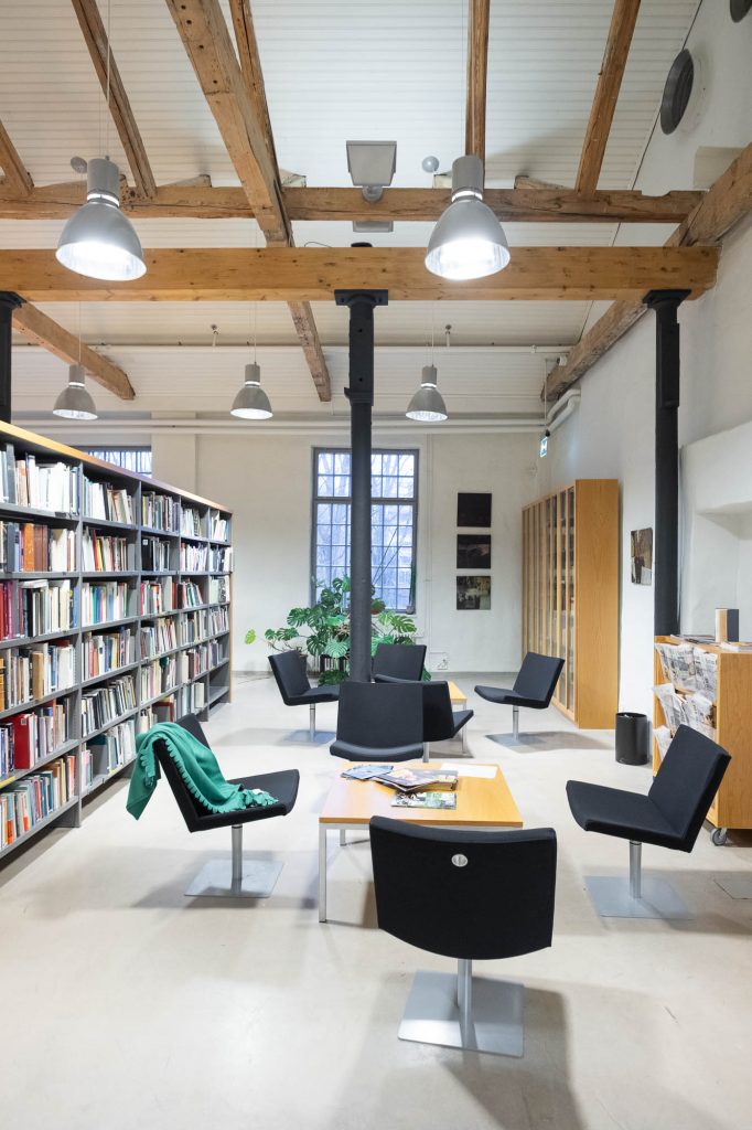 A modern library reading area with black chairs, tables, bookshelves filled with books, and exposed wooden ceiling beams. A green coat is draped over one chair, and large windows let in natural light.